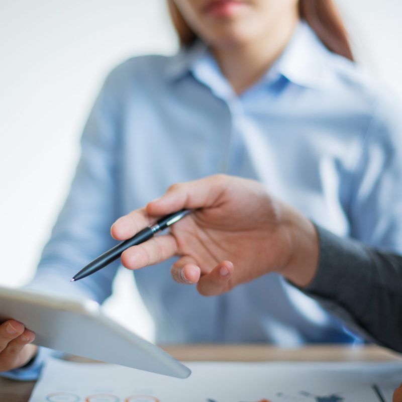 Unrecognizable Asian business team having meeting in office. Businesswoman holding touchpad, businessman pointing at touchscreen with pen and discussing information